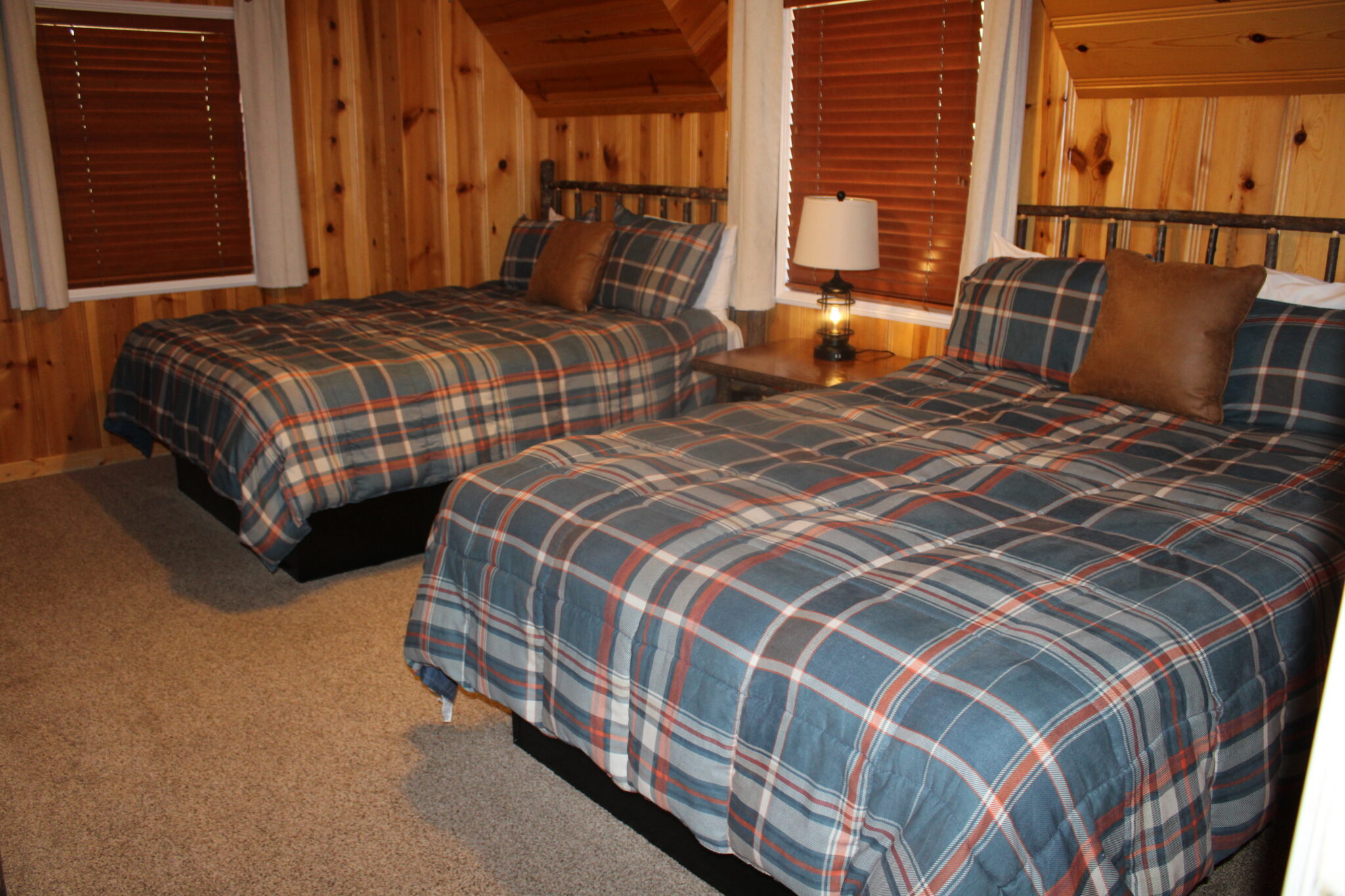 A cozy guest room with two twin beds, plaid bedspreads, wood paneling on the walls, and two windows with blinds.