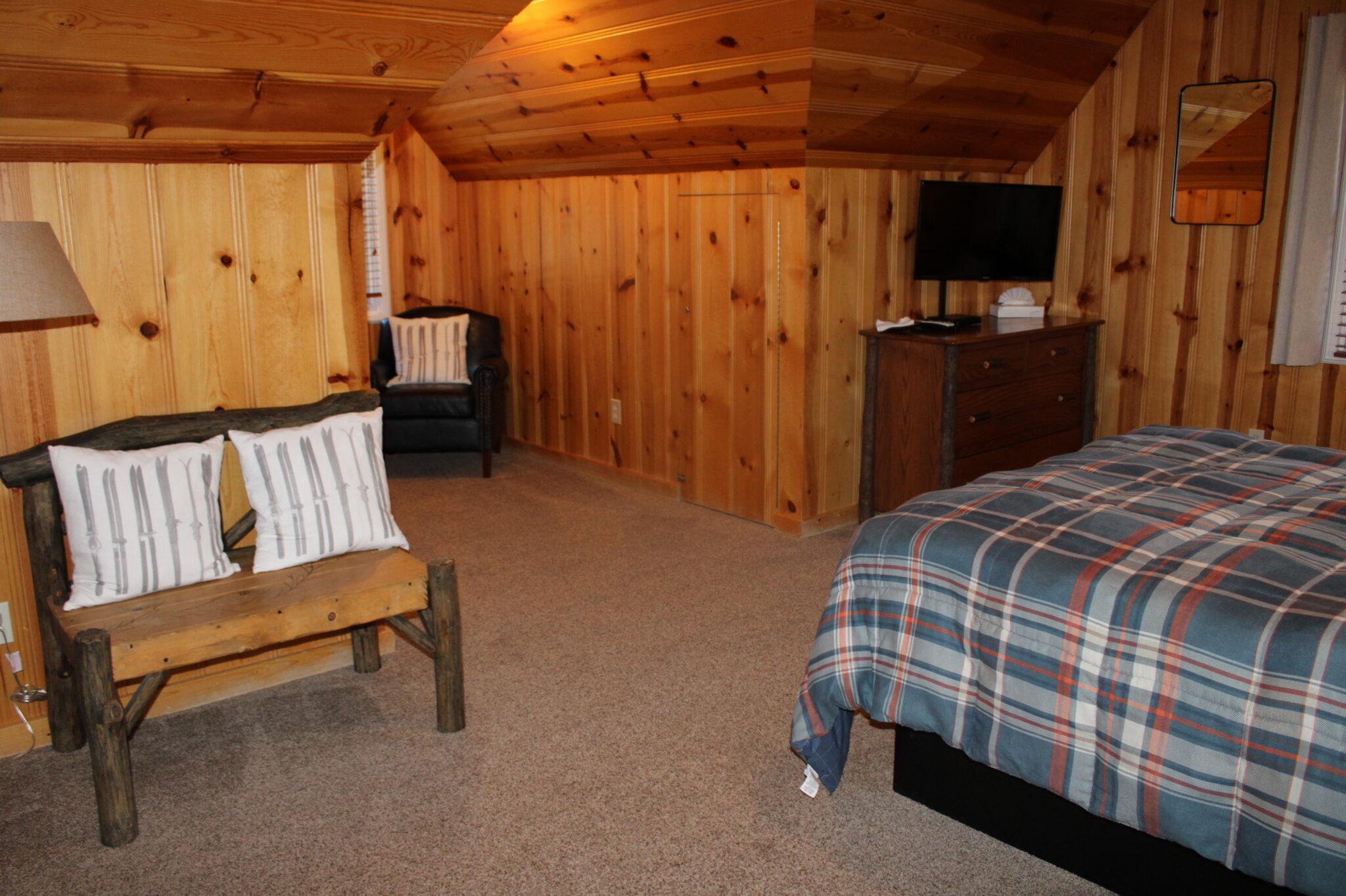 A wide view of a room with wood paneling, a bed with a plaid bedspread, a flat-screen TV on a dresser, an armchair, and a bench.